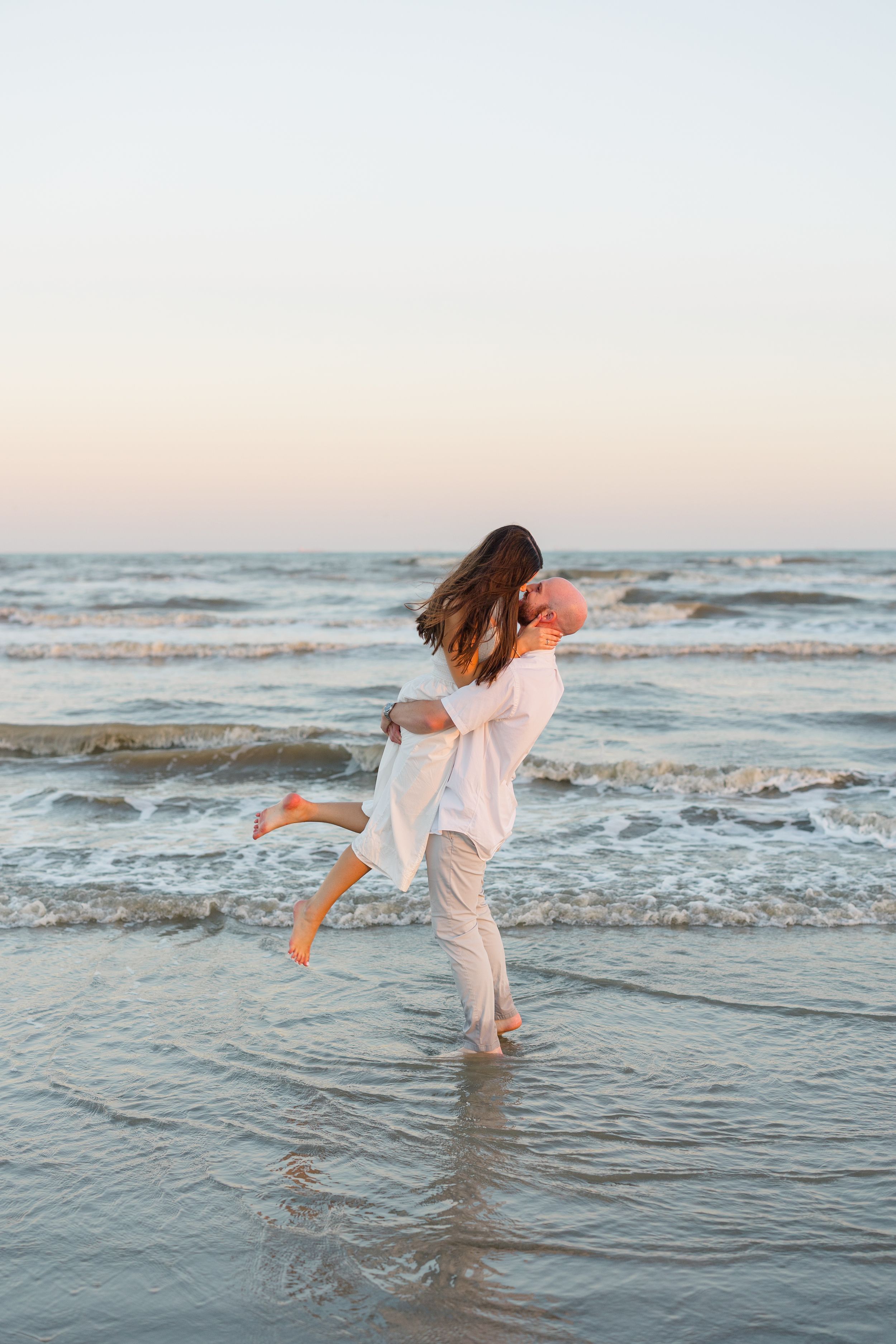 Man lifting woman in white dress at the ocean shoreline during a romantic beach session at sunset.
