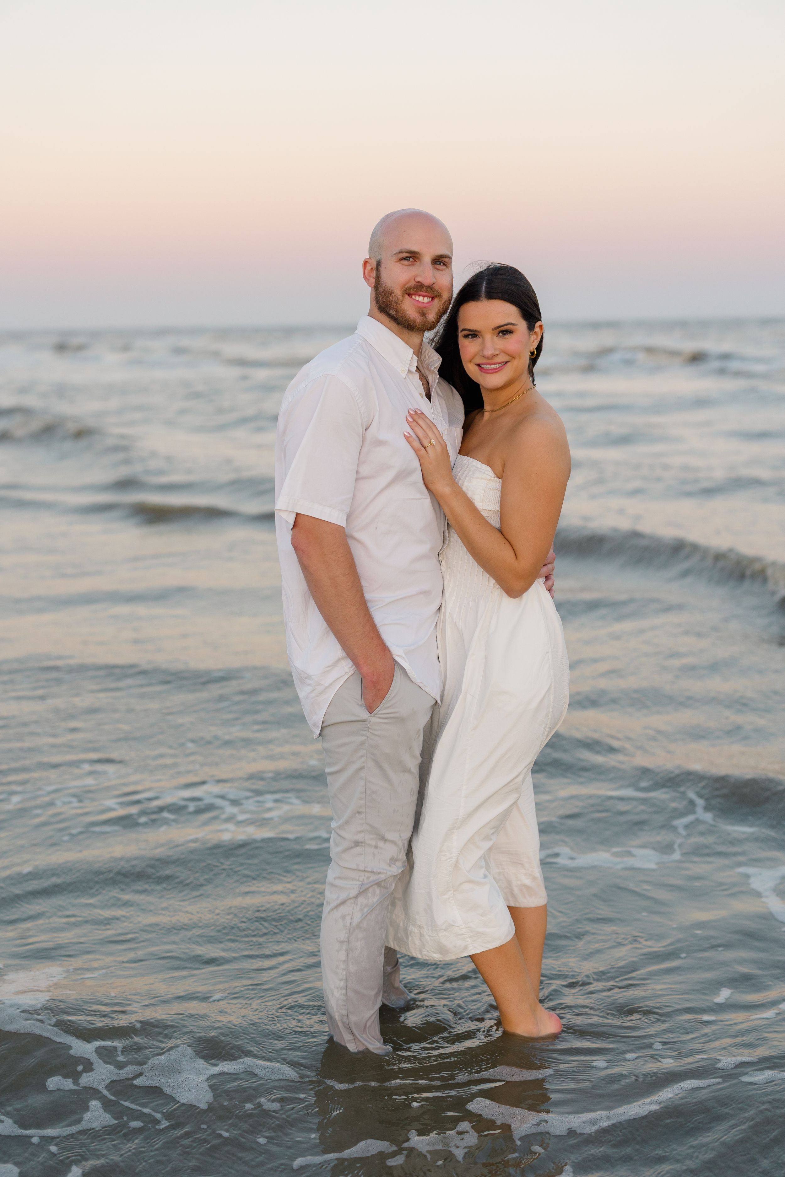 Smiling couple standing in ocean waves at sunset, woman in white jumpsuit embracing bald man in white shirt.