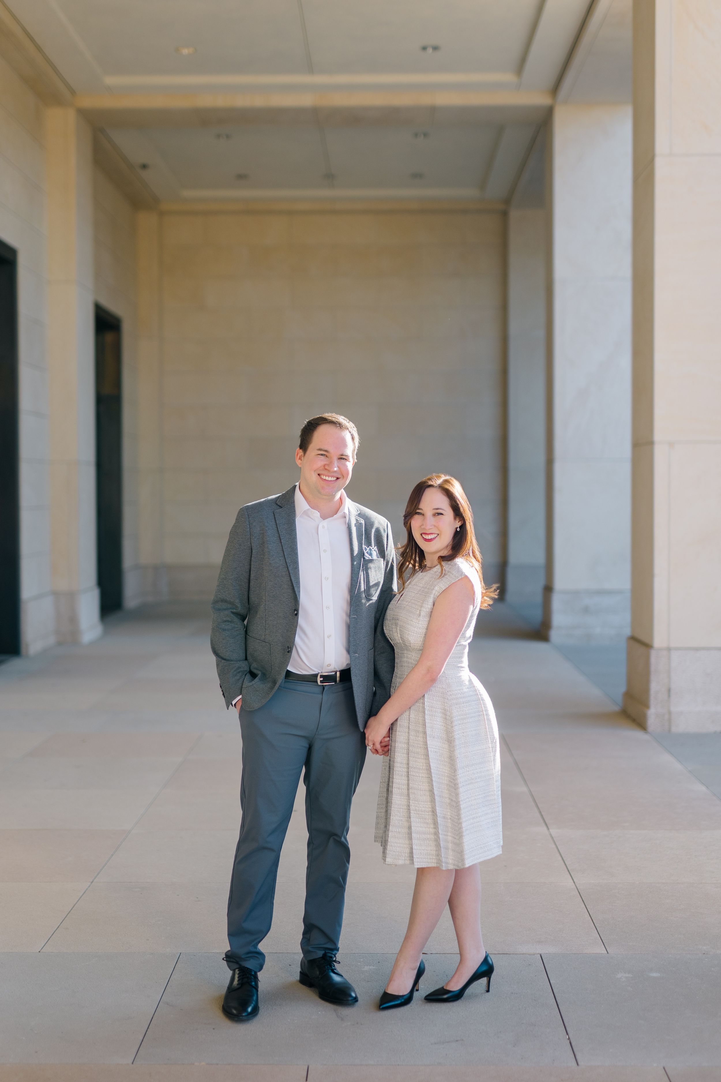 Smiling couple in formal attire posing in sunlit stone corridor for engagement photos.