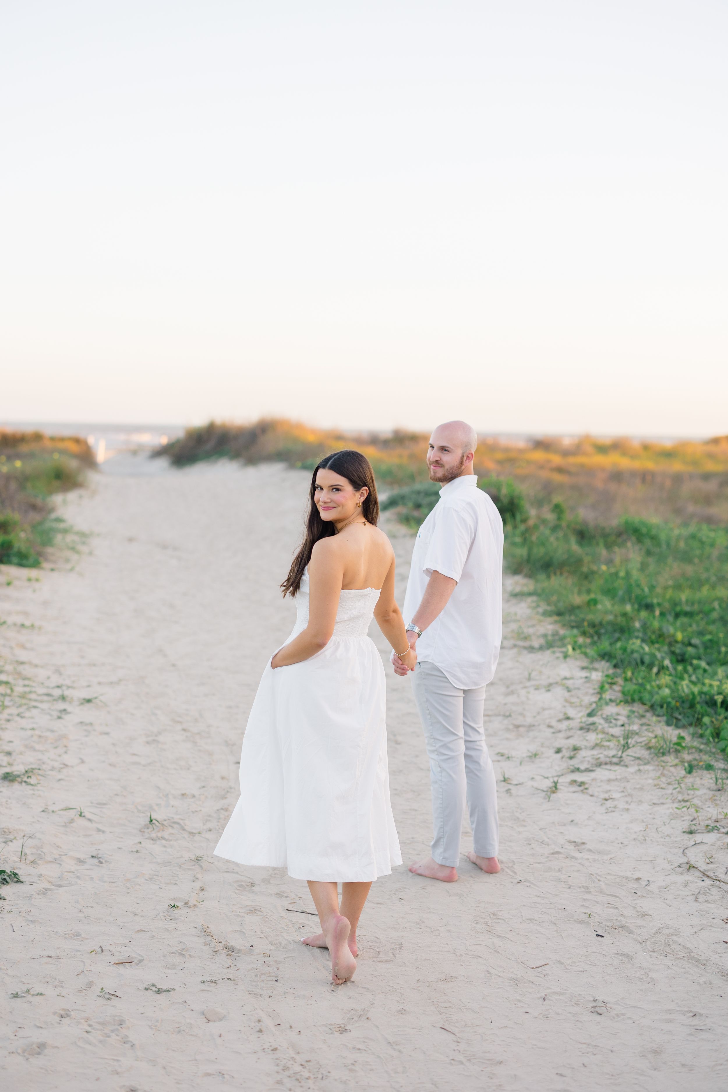 Couple holding hands on sandy beach path at sunset, woman in white strapless dress looking back over shoulder.