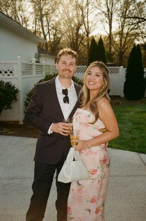 Couple posing outdoors at a garden event, man in dark suit, woman in floral dress, holding drinks at golden hour.
