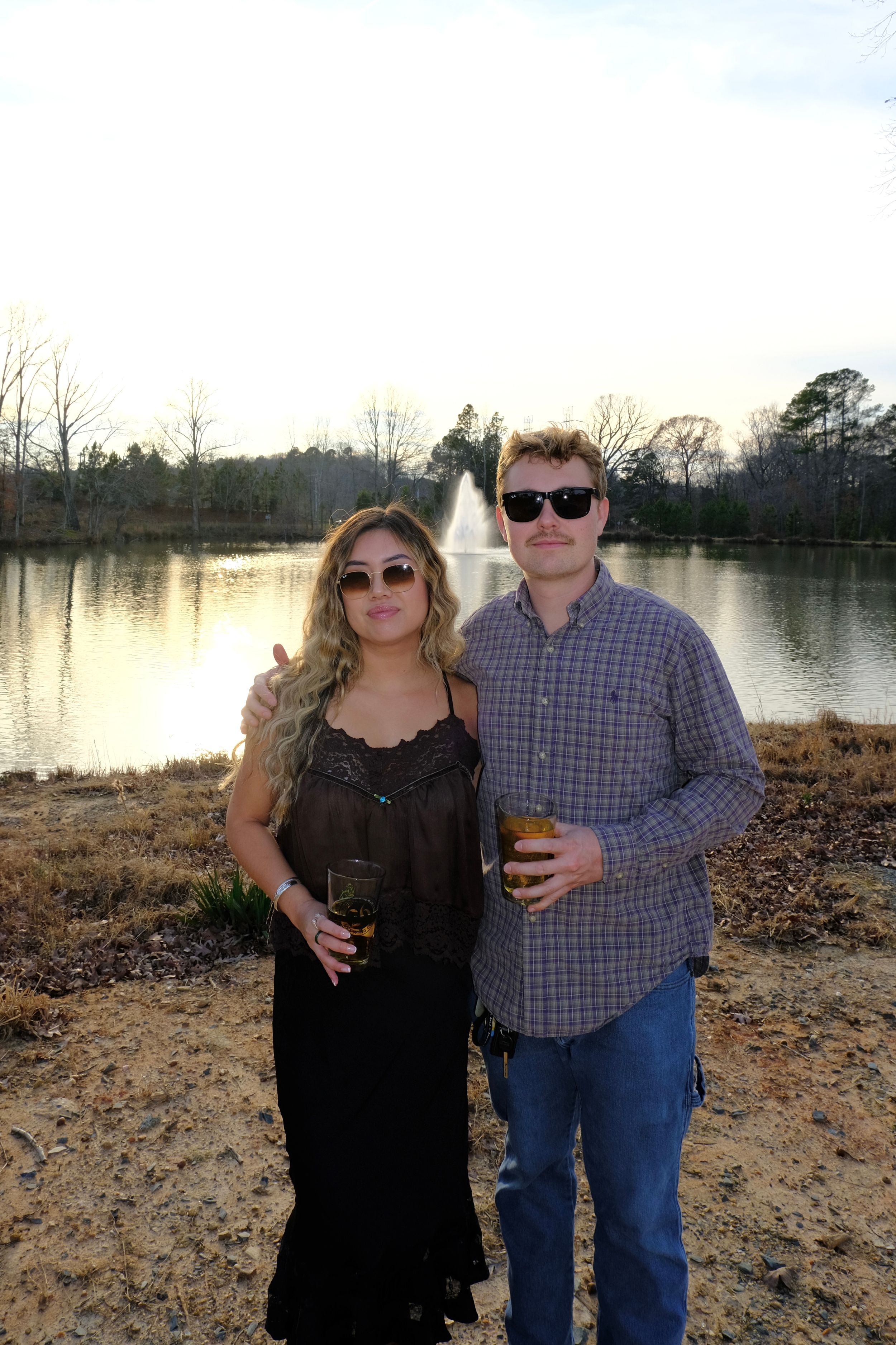 Couple posing by a serene pond with a fountain, woman in black dress holding wine, man in plaid shirt with sunglasses.