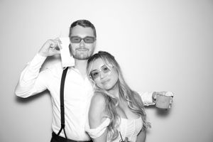Black and white photo booth picture of a smiling couple posing together, man in suspenders and woman in white dress.