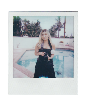 A young blonde woman in a black dress holds a camera poolside with palm trees in the background.