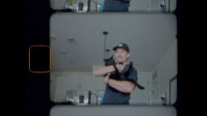 Man in black cap holds a small black cat up in a home kitchen, captured in vintage 8mm film style.