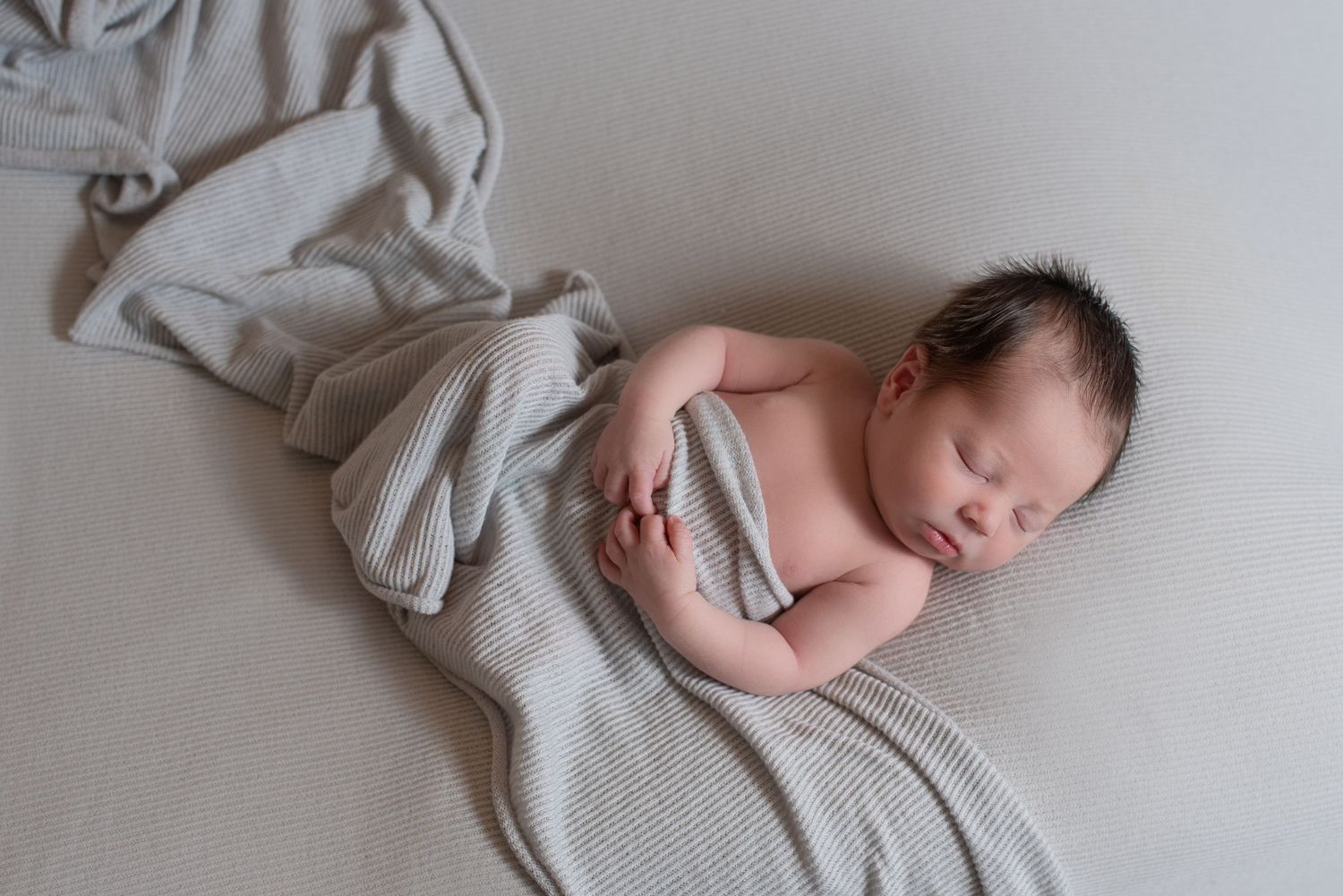 Peaceful sleeping newborn partially wrapped in soft gray blanket on white surface.