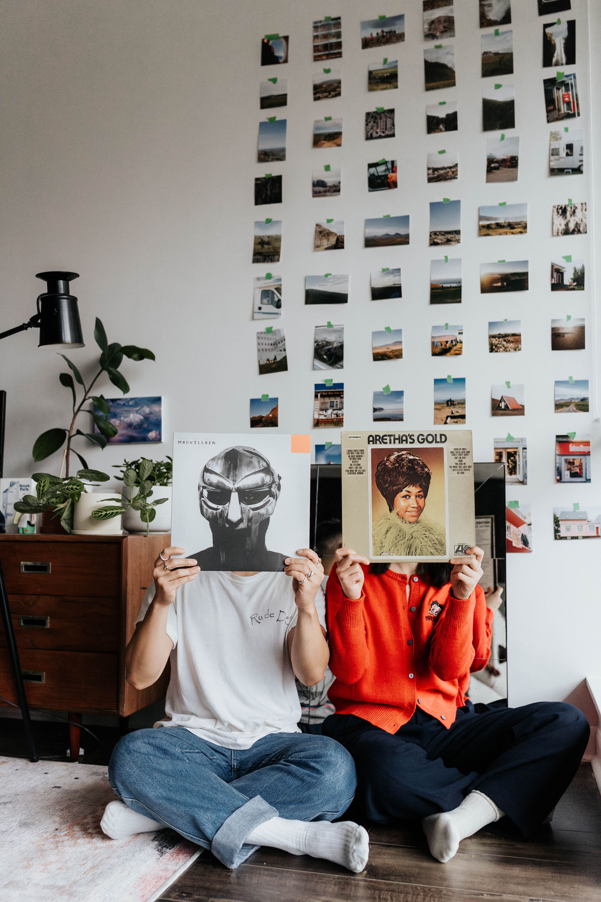 Two people holding vinyl records in front of their faces with a photo wall display behind them.