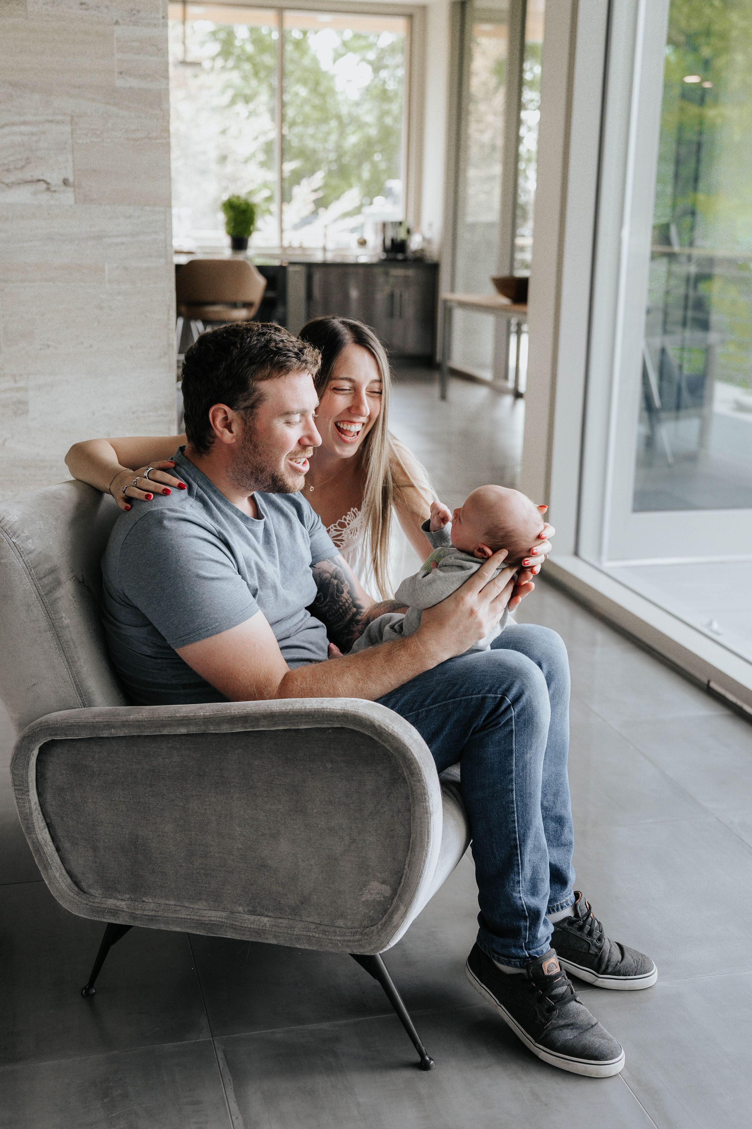 A couple sits together on a gray couch in a bright room with large windows, sharing a tender moment.