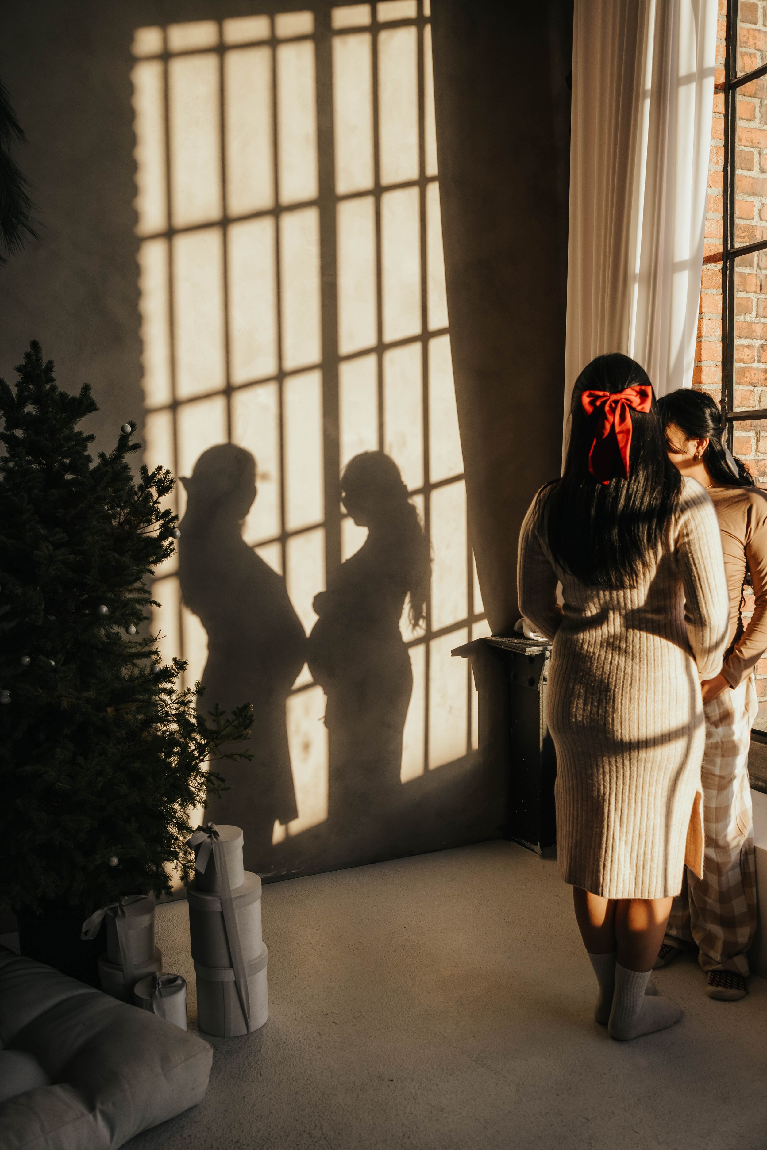 Dramatic shadows cast on wall from sunlight through window as figure stands in cream knit dress by Christmas tree.