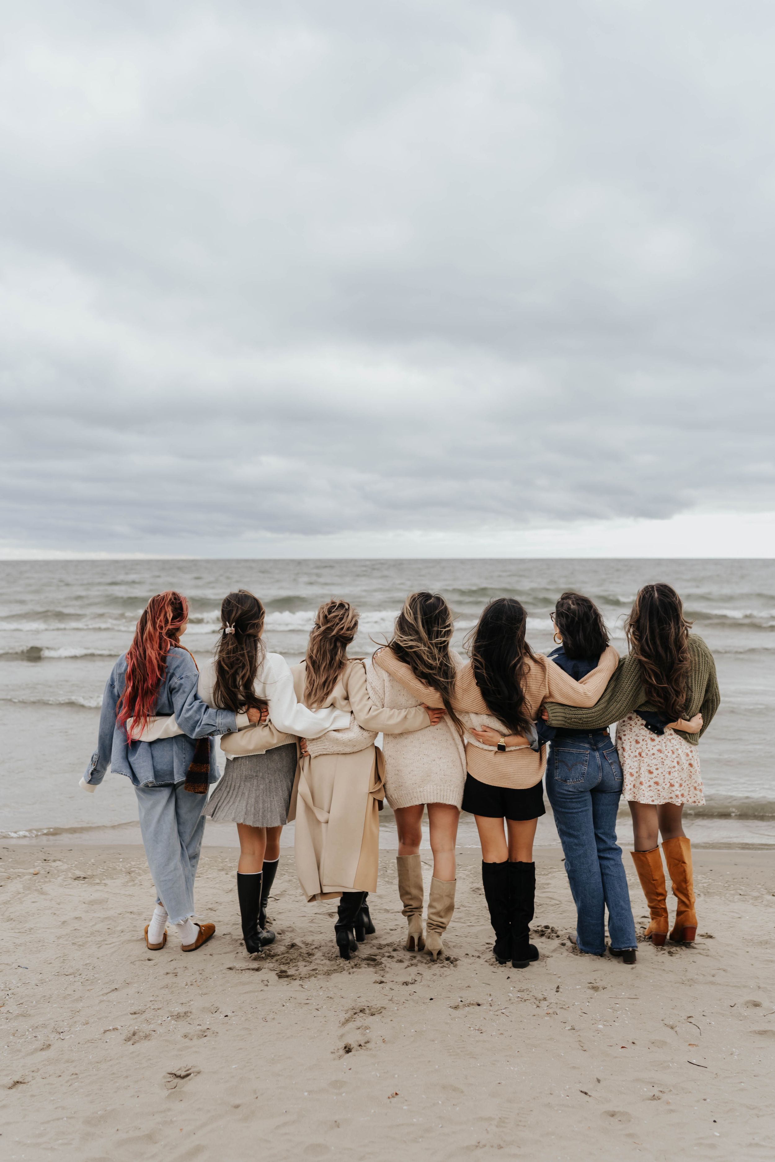 A group of friends stand with their backs to camera in a line along a cloudy beach shoreline.