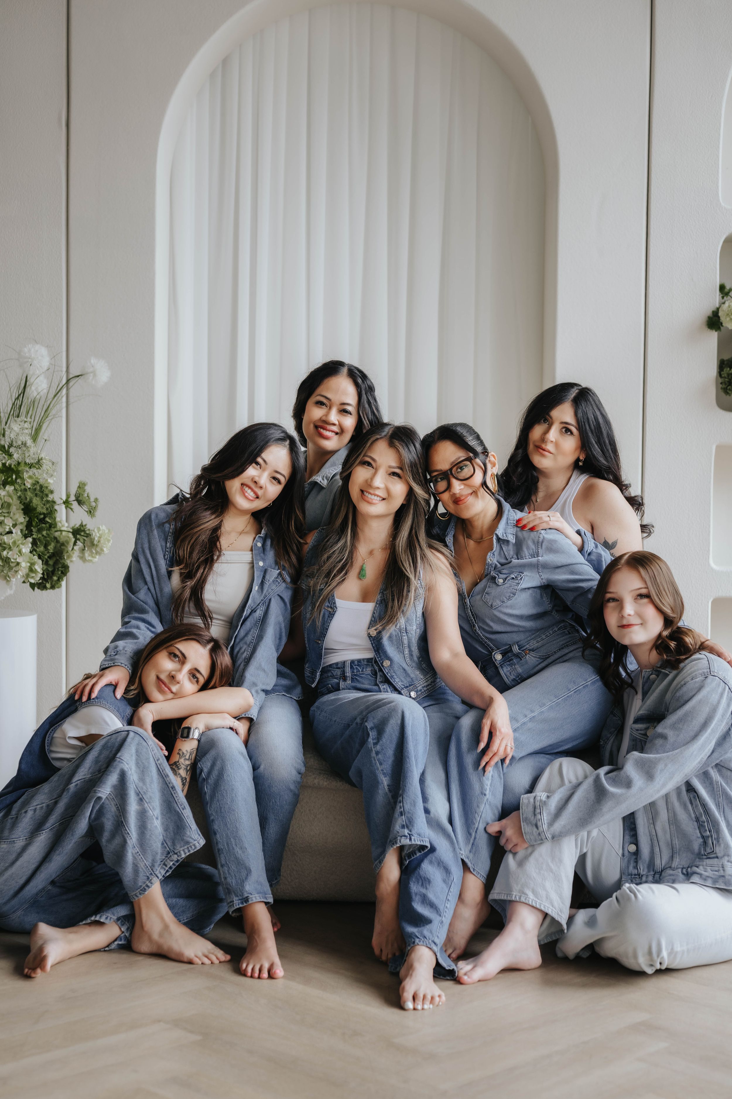 Six women in matching denim outfits pose together smiling in a bright white studio setting.
