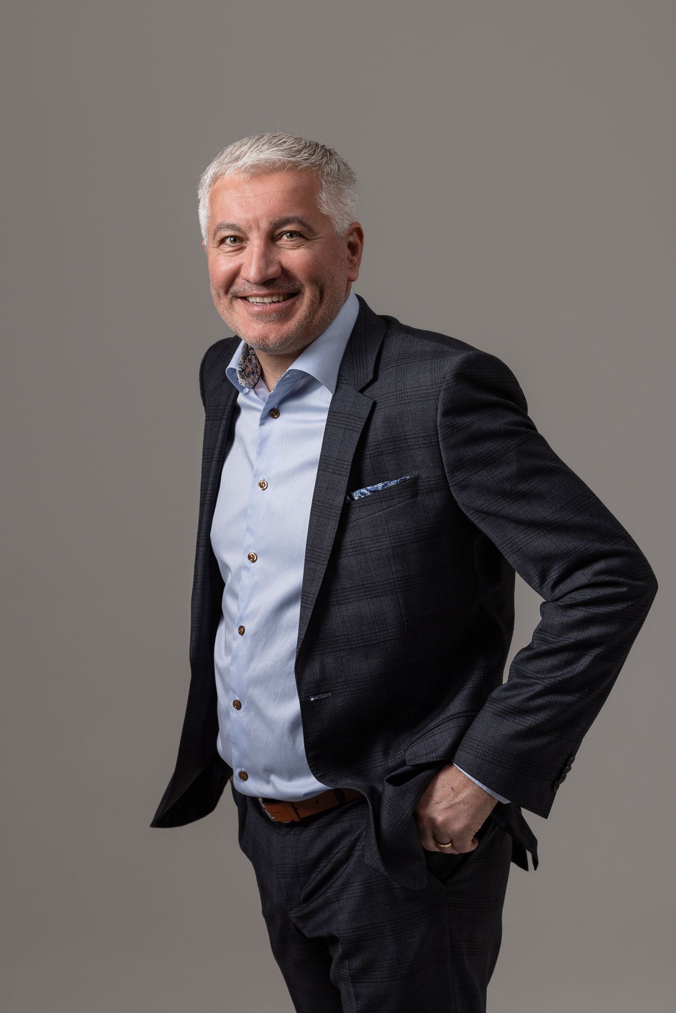 Professional studio portrait of someone in a dark suit jacket and light blue dress shirt posing against a gray background.