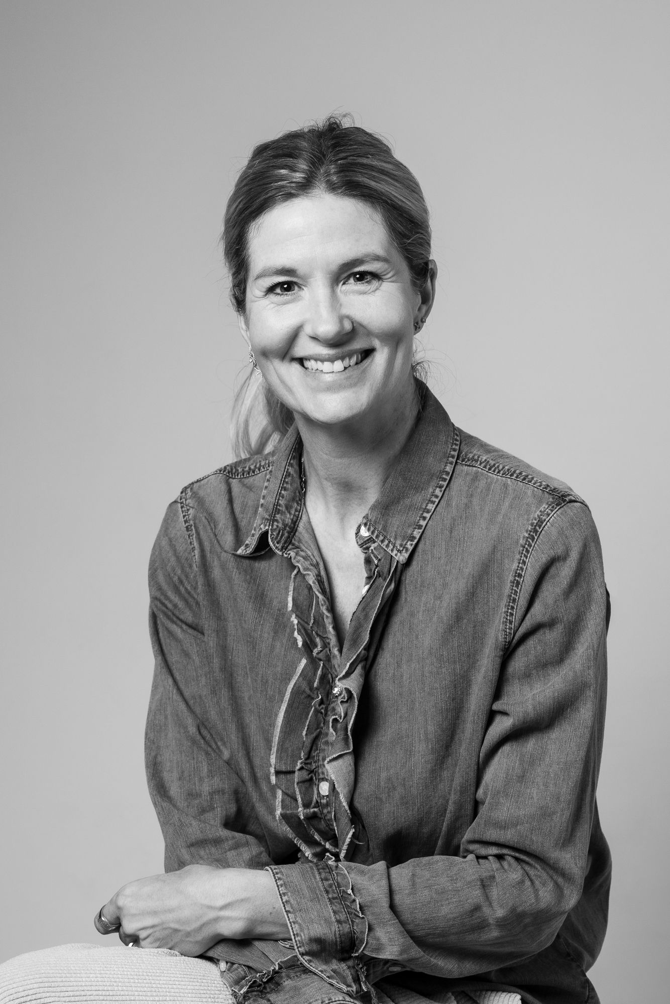 A black and white studio portrait of someone wearing a casual denim shirt while smiling warmly at the camera.