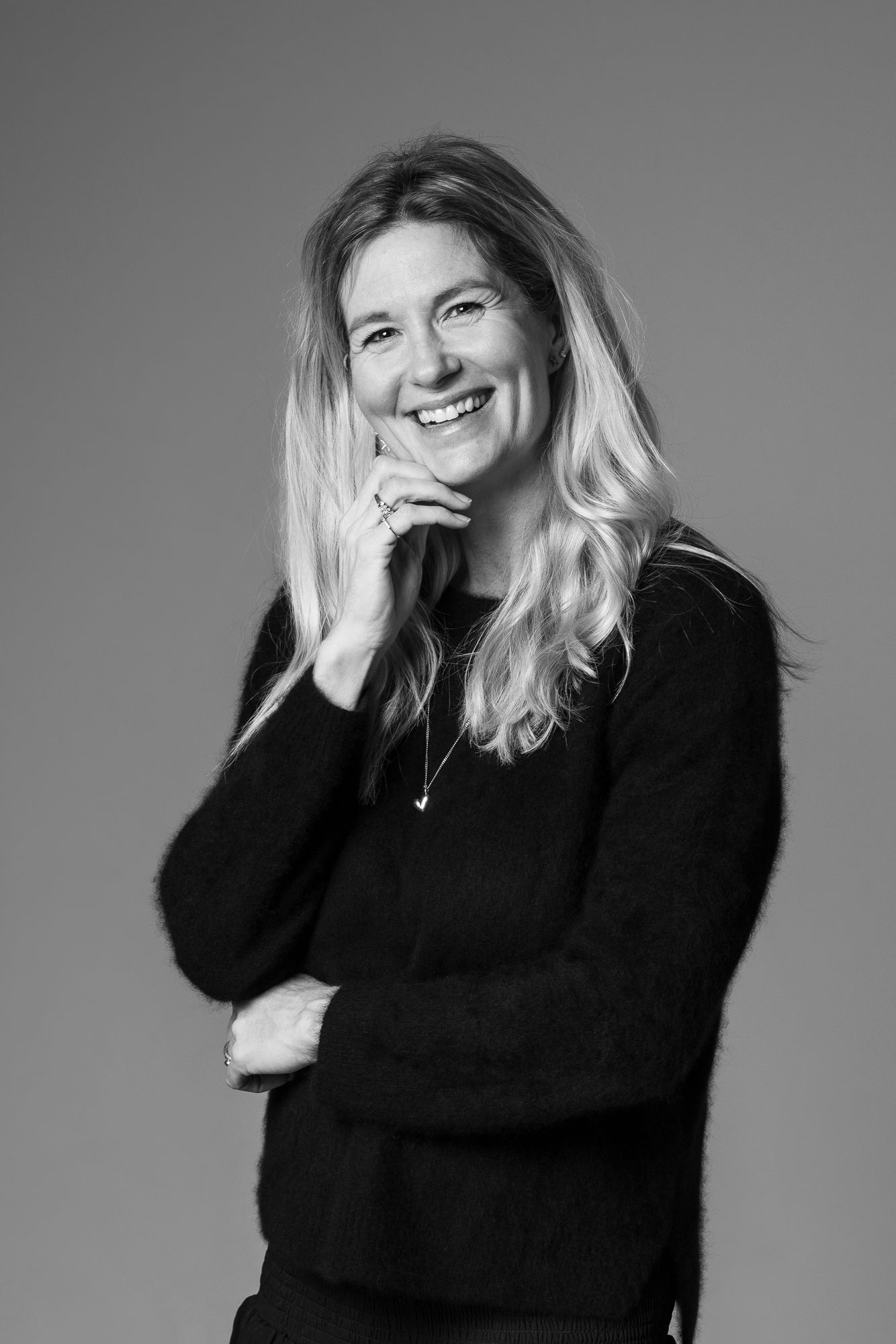 A black and white portrait photograph shows a person wearing a black sweater smiling warmly against a grey background.