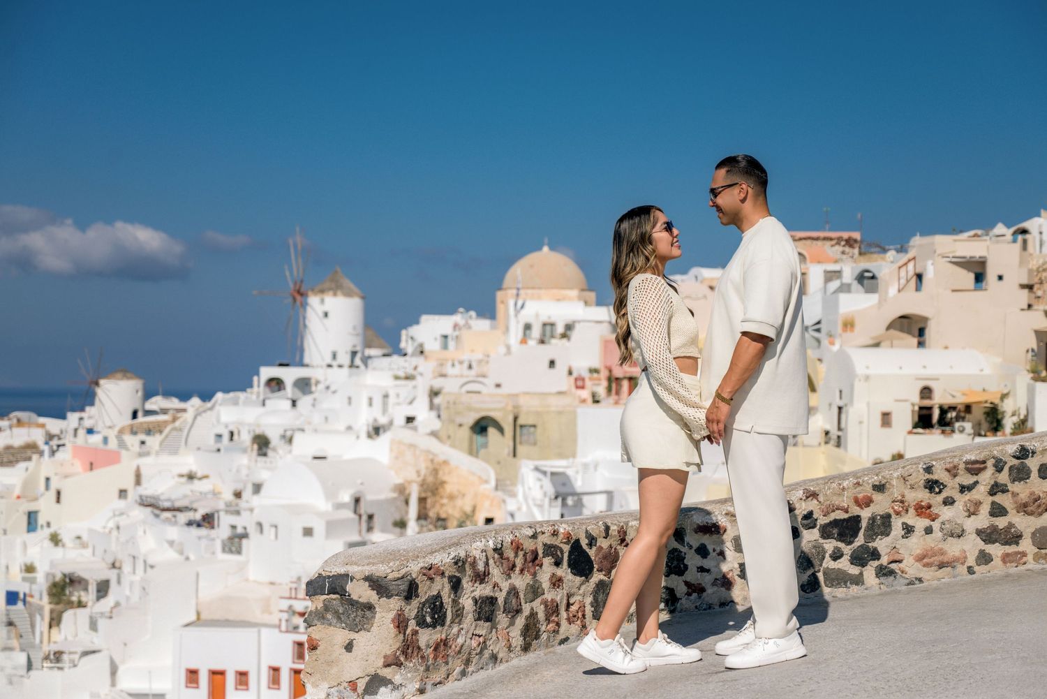 A couple in white outfits enjoying a romantic moment in Santorini, Greece with iconic whitewashed buildings and windmill background.