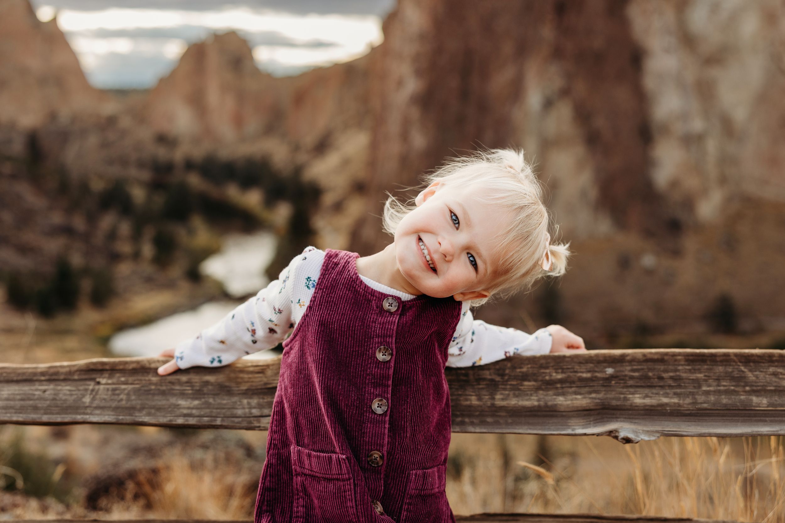 A child in a burgundy corduroy dress leans playfully on a wooden fence with red rock formations in the background.