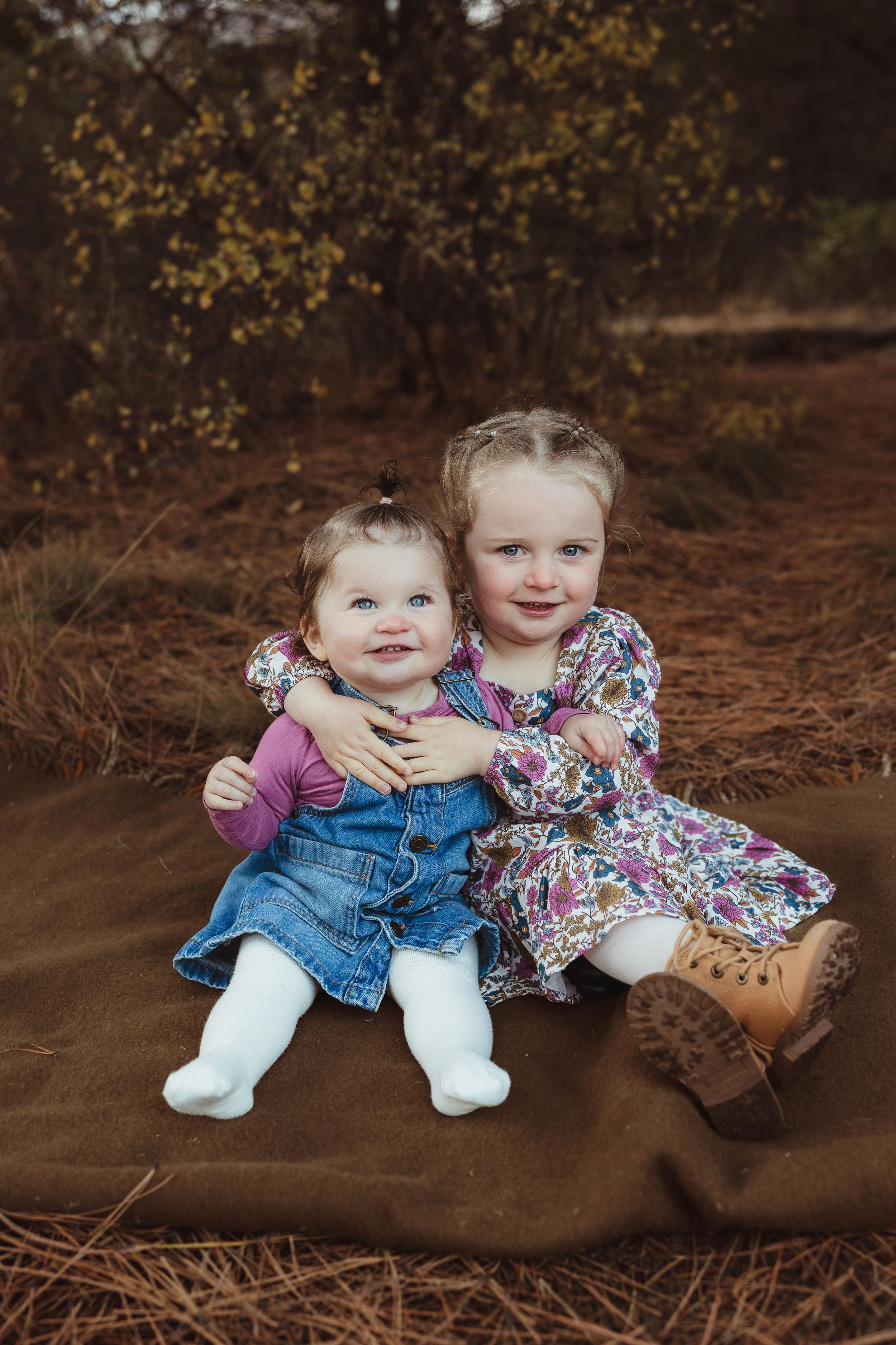 Two children sit together on a fallen log in an autumn forest setting wearing denim and floral prints.