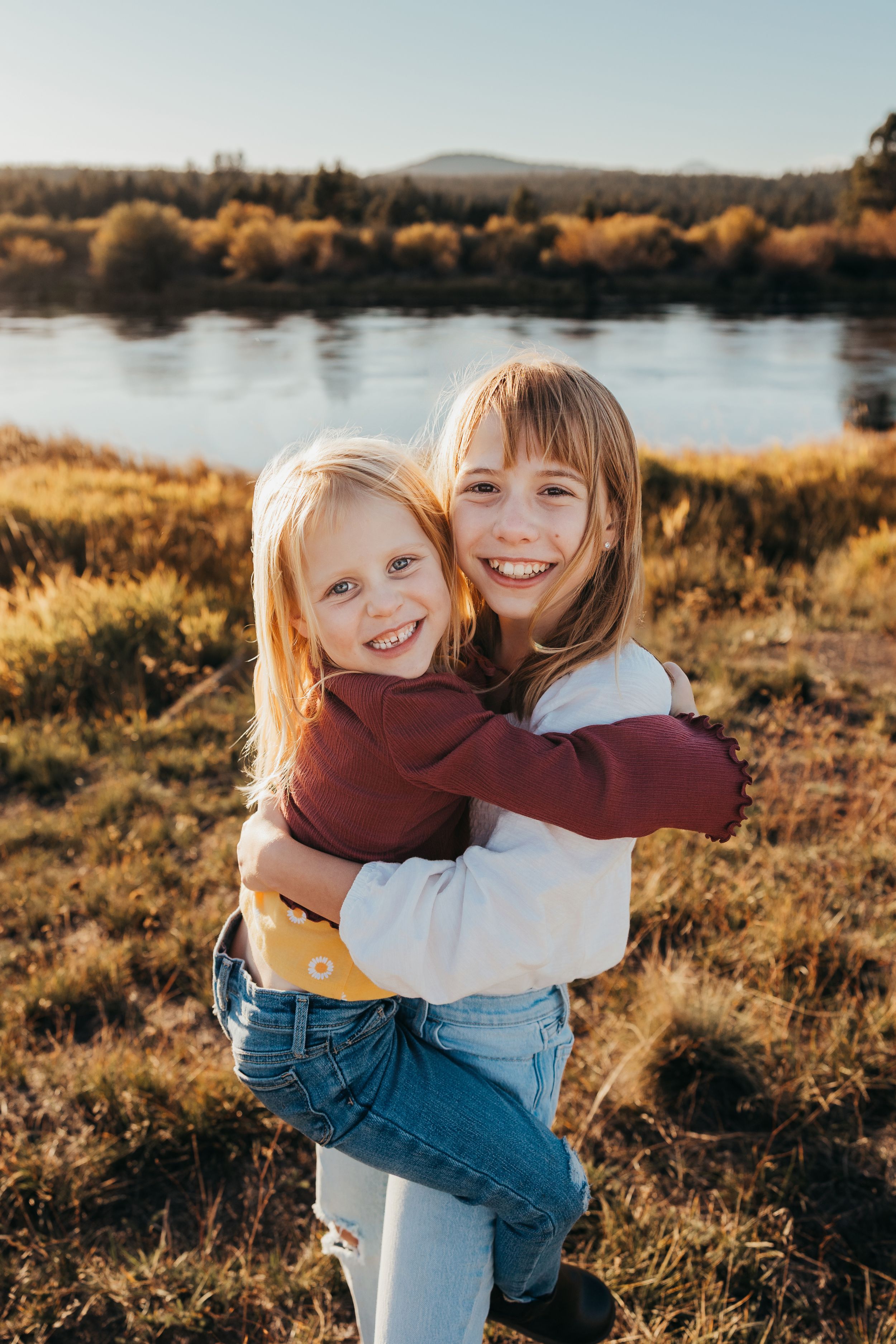 Two people embrace while standing in a golden field at sunset near a lake.