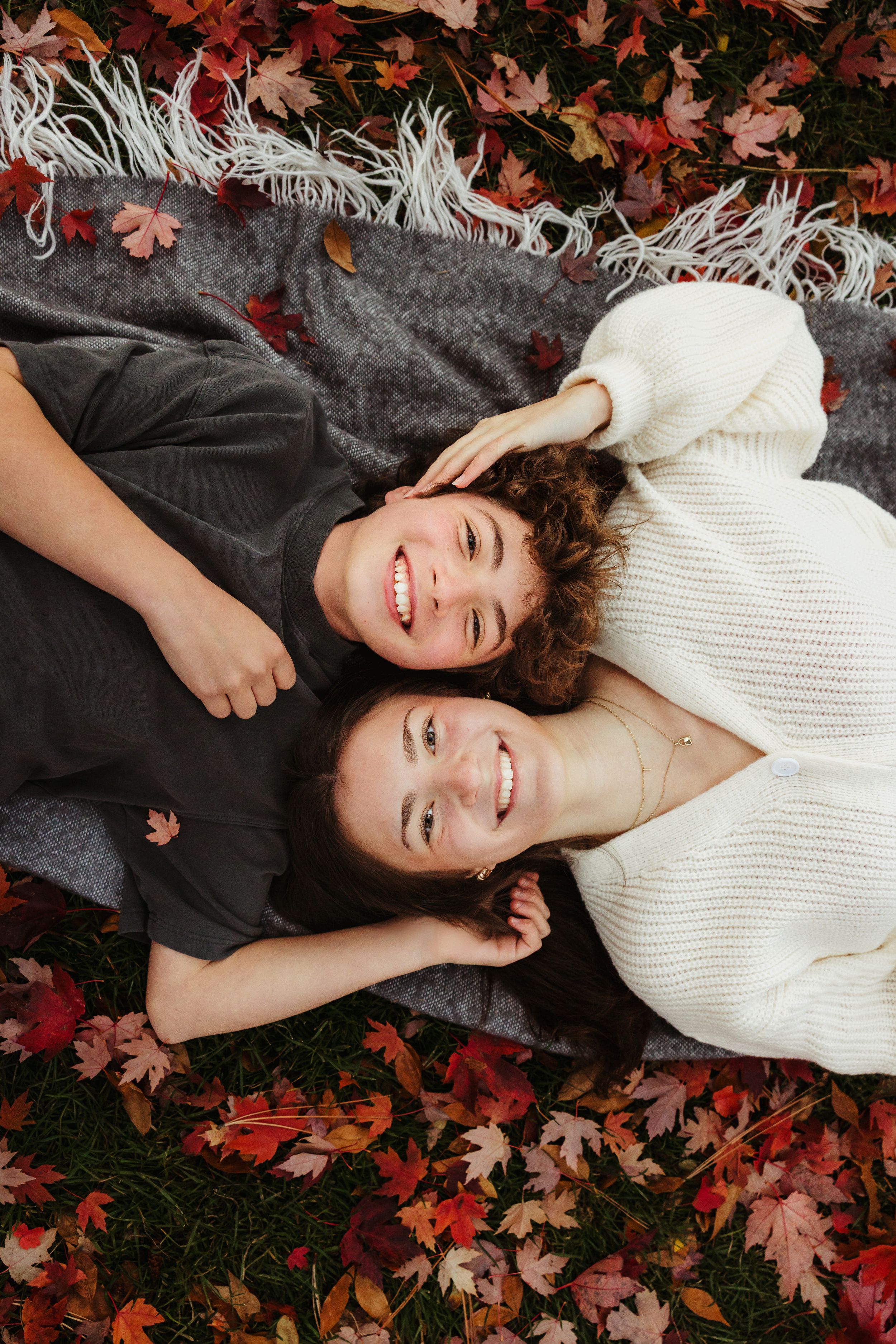 Two people lying on a gray blanket surrounded by red autumn leaves while smiling and wearing cozy sweaters.