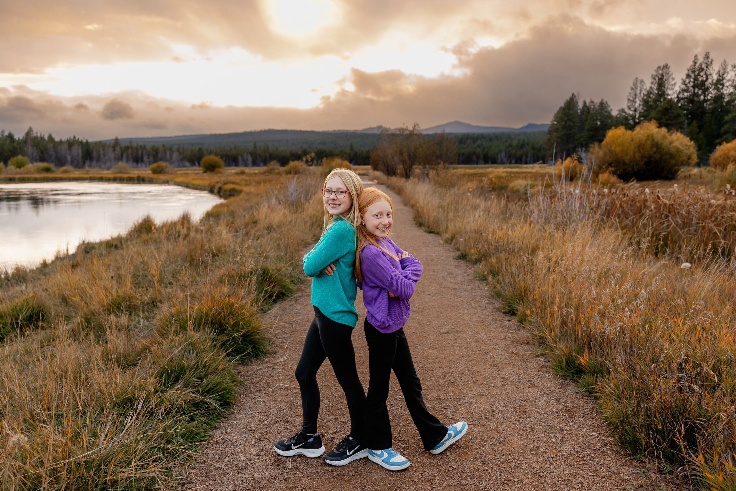 A group of people walking along a scenic mountain trail at sunset with tall grass and mountains in the background.