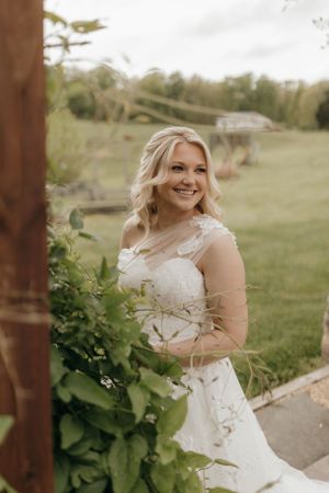 Bride in elegant lace wedding dress poses by greenery outdoors.