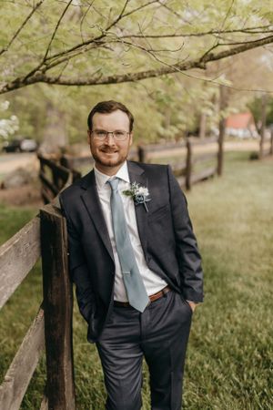 Groom in classic dark suit poses by wooden fence under spring branches.