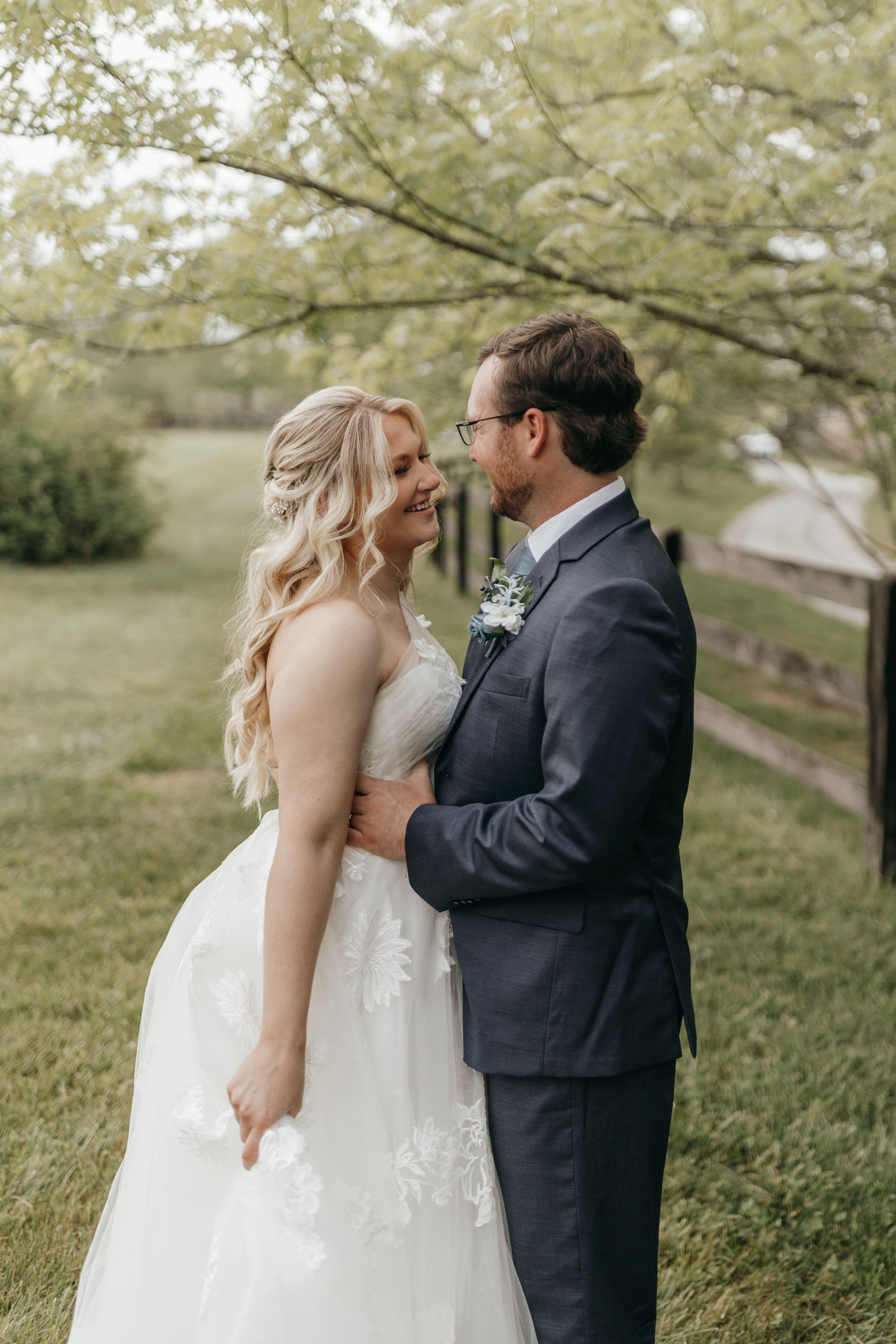 Beautiful outdoor wedding portrait featuring bride in lace gown and groom in gray suit under autumn trees.