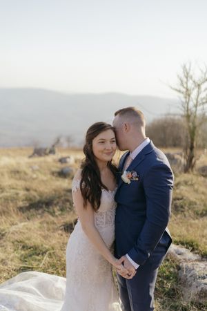 A couple in wedding attire embrace on a hilltop during golden hour with mountains in the background.