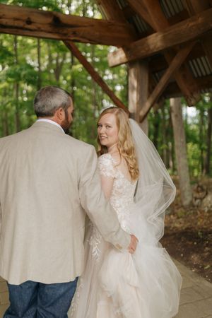 A bride and her father share a tender first look moment under a rustic wooden pavilion in a forest setting.