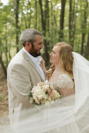 A romantic wedding couple embraces in a sunlit forest setting while the bride's veil flows in the breeze.