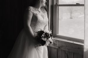 A bride in a white lace wedding dress stands near a window, creating a soft silhouette in natural light.