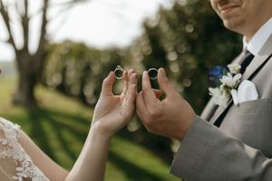 Close-up shot of hands holding wedding rings during an outdoor ceremony with bokeh background and white boutonniere.
