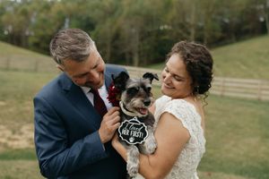 A couple in wedding attire share a tender moment with their dog in an outdoor field setting.