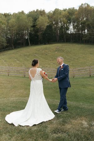 A couple in wedding attire dances playfully in a grassy meadow with trees in the background.