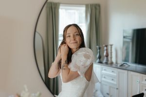 A bride in a white wedding dress stands in front of a window while getting ready for her special day.