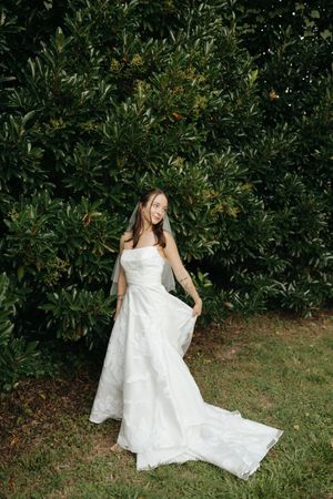 A person in a flowing white dress poses gracefully against a backdrop of lush green foliage in an outdoor garden setting.