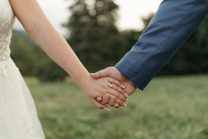 A close-up shot of a bride and groom holding hands, showing wedding rings against a blurred natural background.