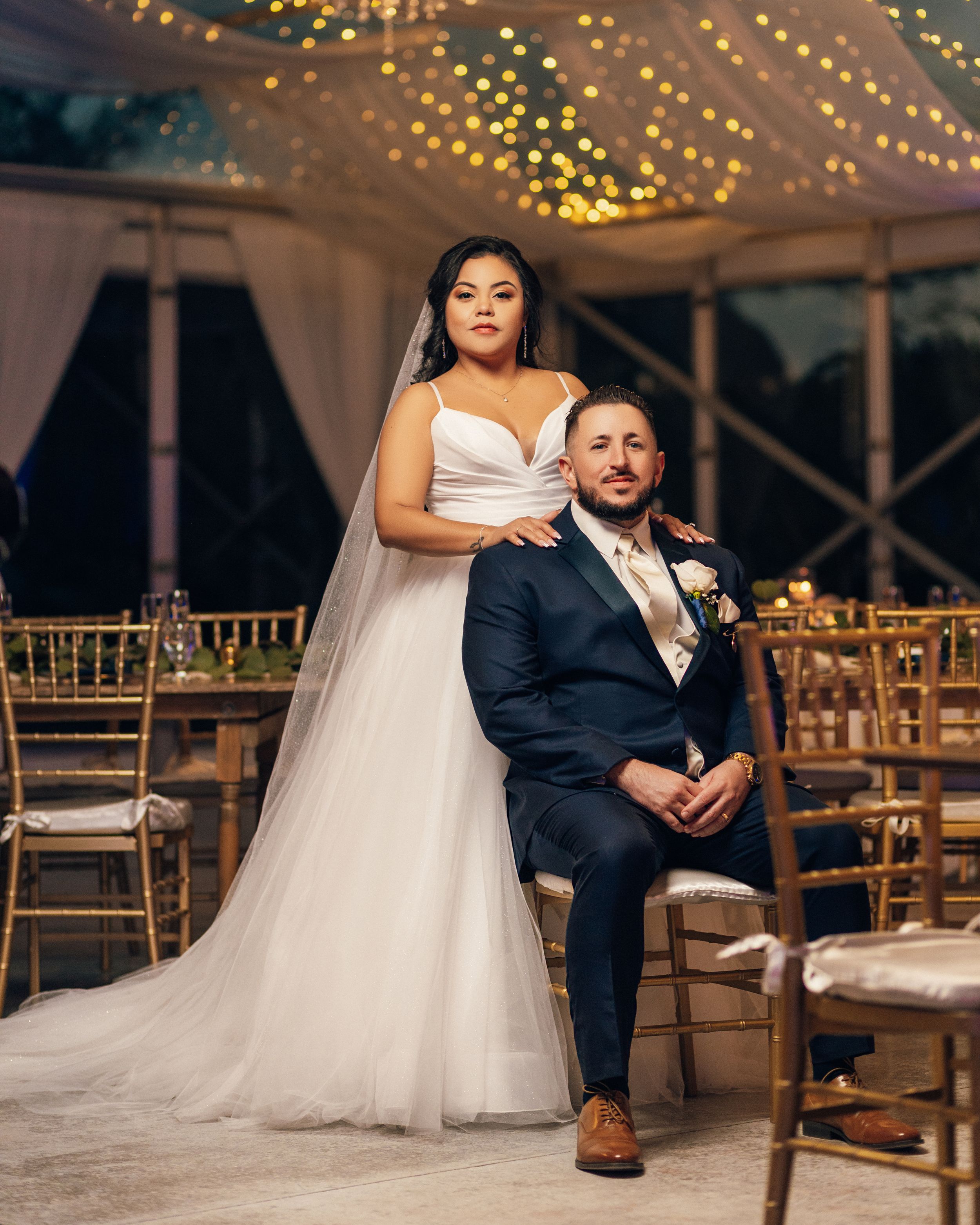 Wedding portrait with bride in white gown and groom seated in reception venue with twinkle lights.