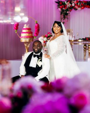 A wedding celebration in a luxurious venue with pink florals, white drapery and an elegant tiered cake display in the background.