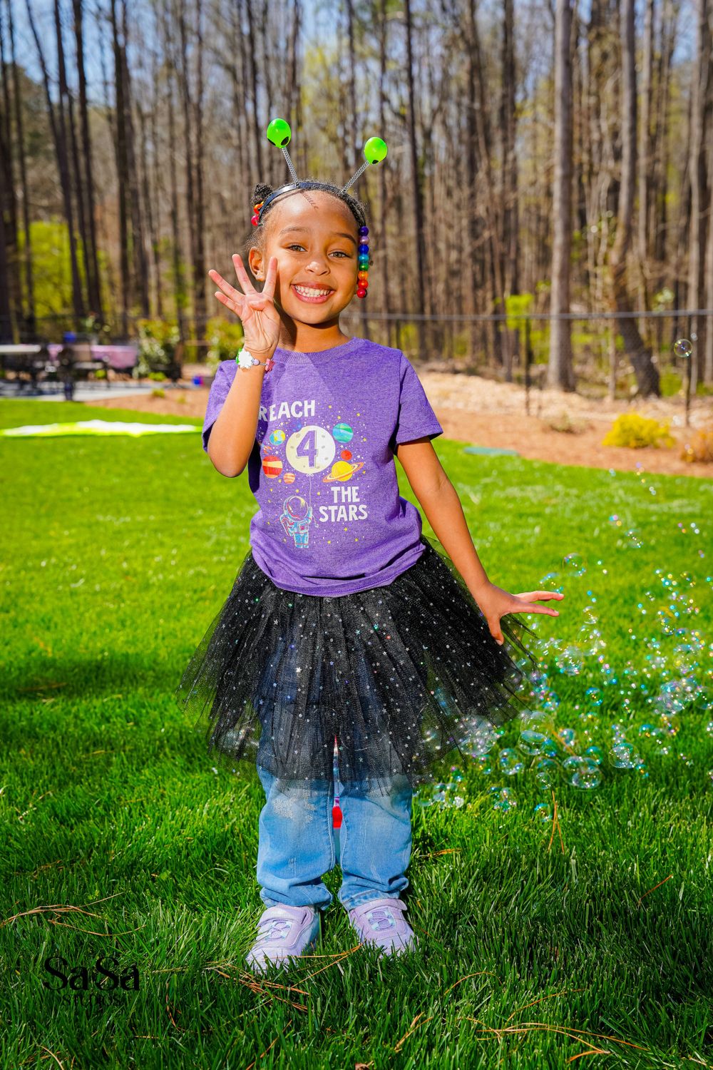 A small child poses outdoors wearing a purple t-shirt, tulle skirt over jeans, and green hairbows in a wooded backyard setting.