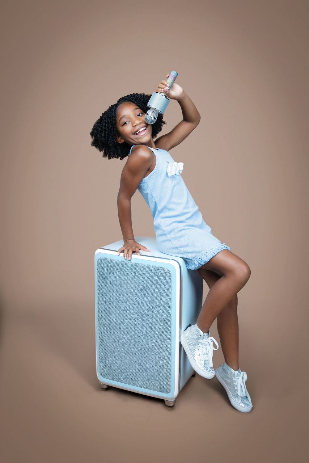 Smiling young Black girl in blue dress sitting on light blue suitcase holding microphone on beige background.