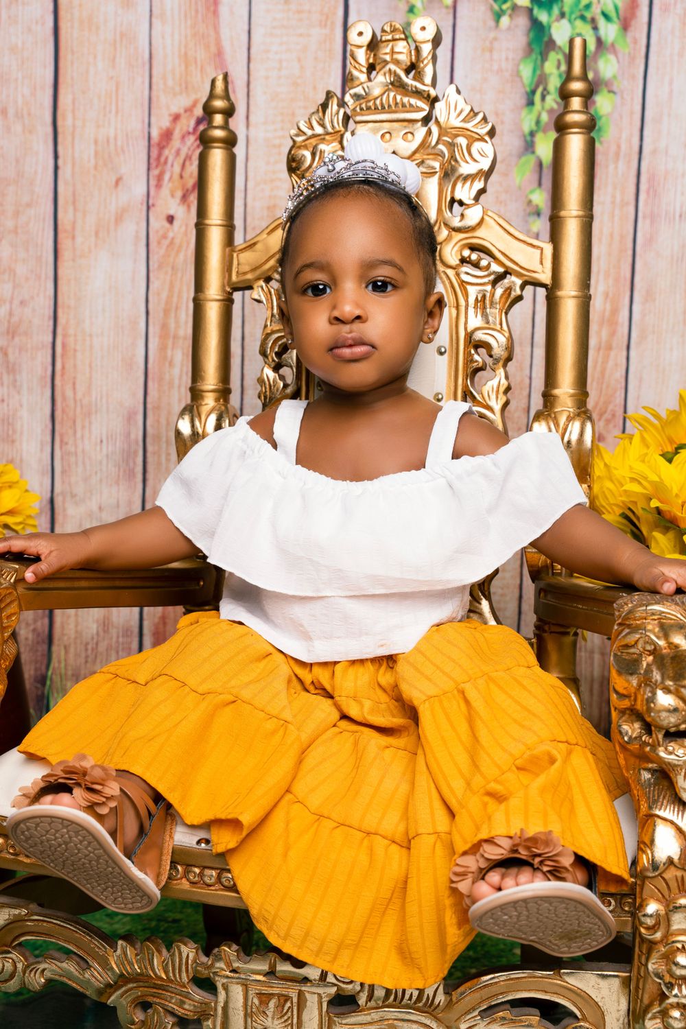 Small child in yellow dress sitting on ornate gold throne with sunflower decorations.