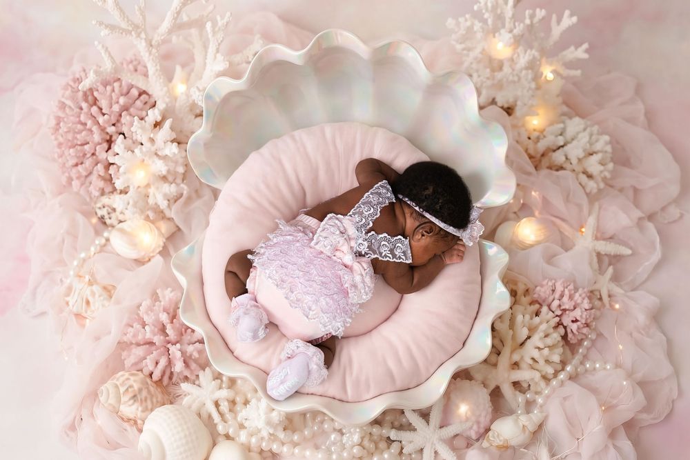 Newborn baby sleeping in a decorative shell bowl surrounded by pink coral and pearls in a dreamy mermaid-themed photo.