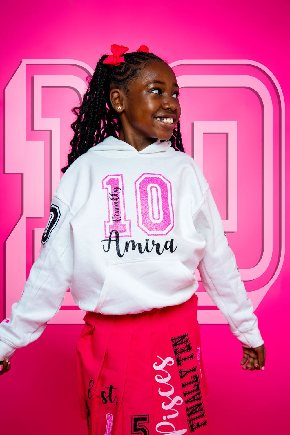 Young dancer wearing a customized white hoodie with 'Amira 10' and pink sweatpants poses against a bright pink backdrop.