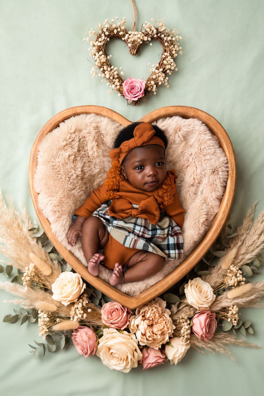 Newborn baby girl in rust outfit resting in heart-shaped wooden bowl with flowers and heart wreath on green backdrop.