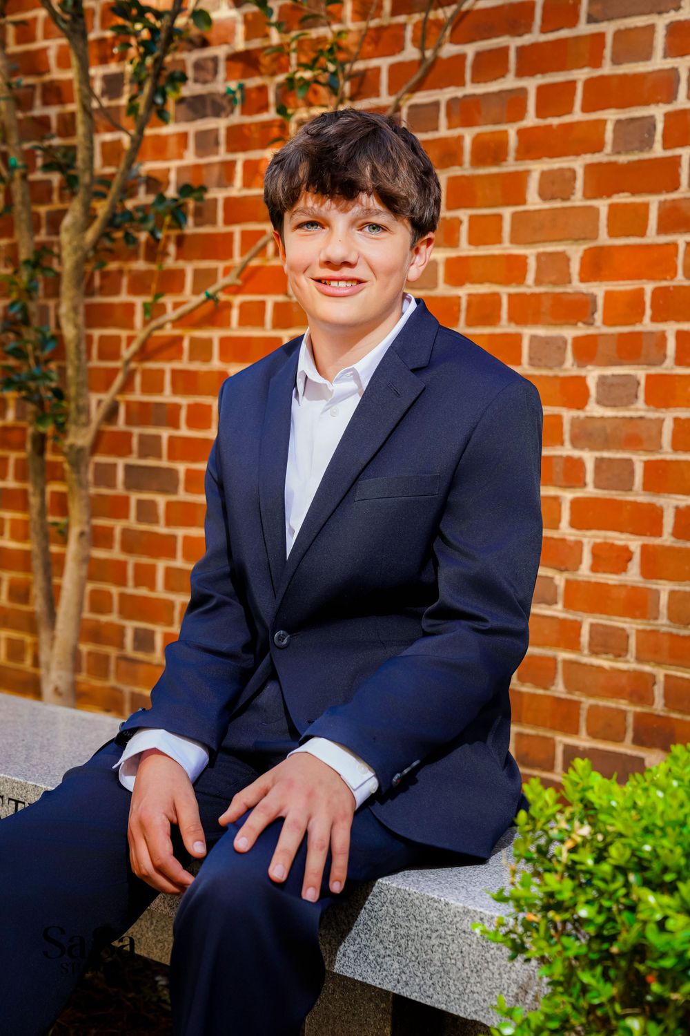 A person wearing a navy blue suit and white shirt sits on a stone bench against a red brick wall.