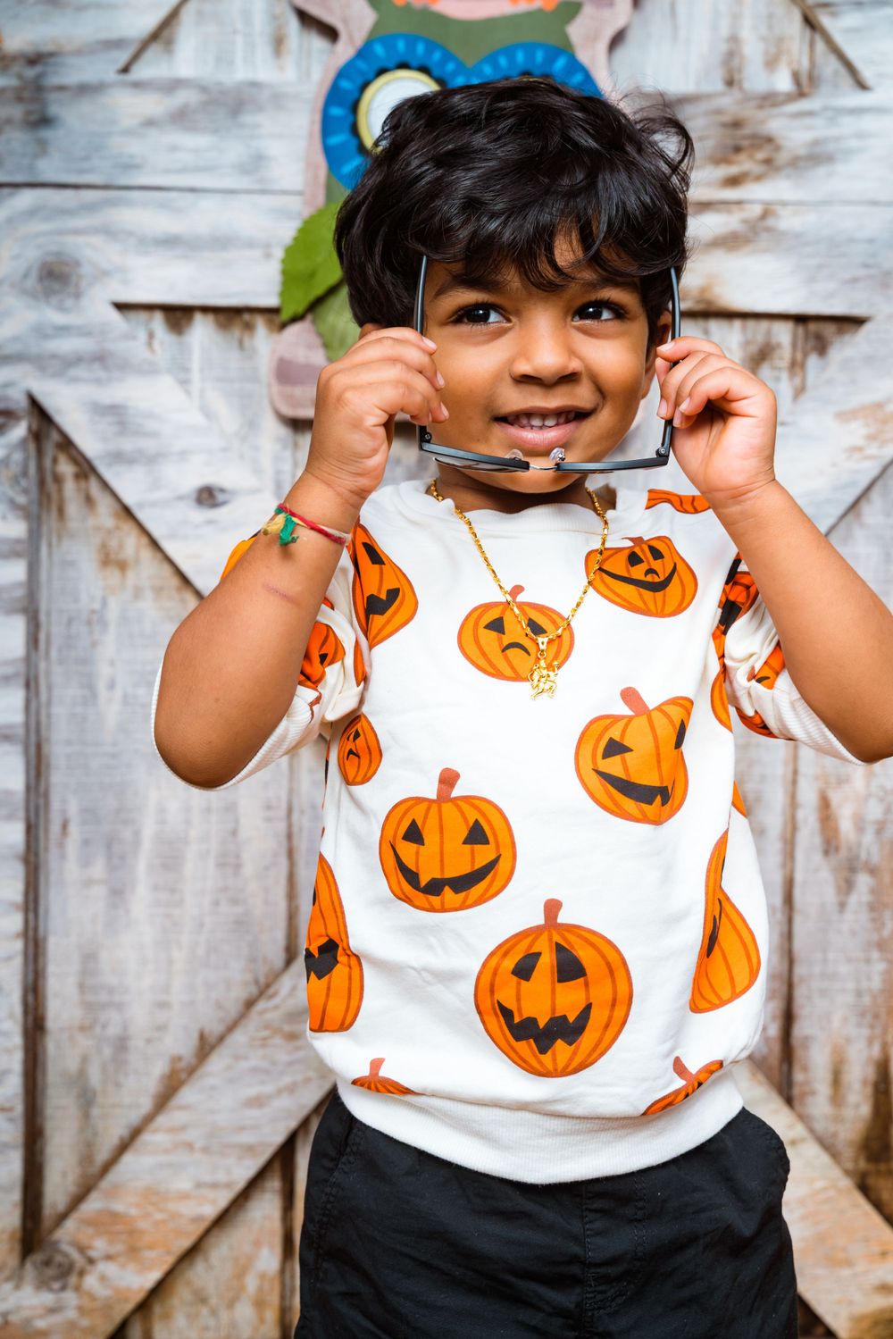 Child wearing pumpkin print shirt poses playfully against rustic backdrop.