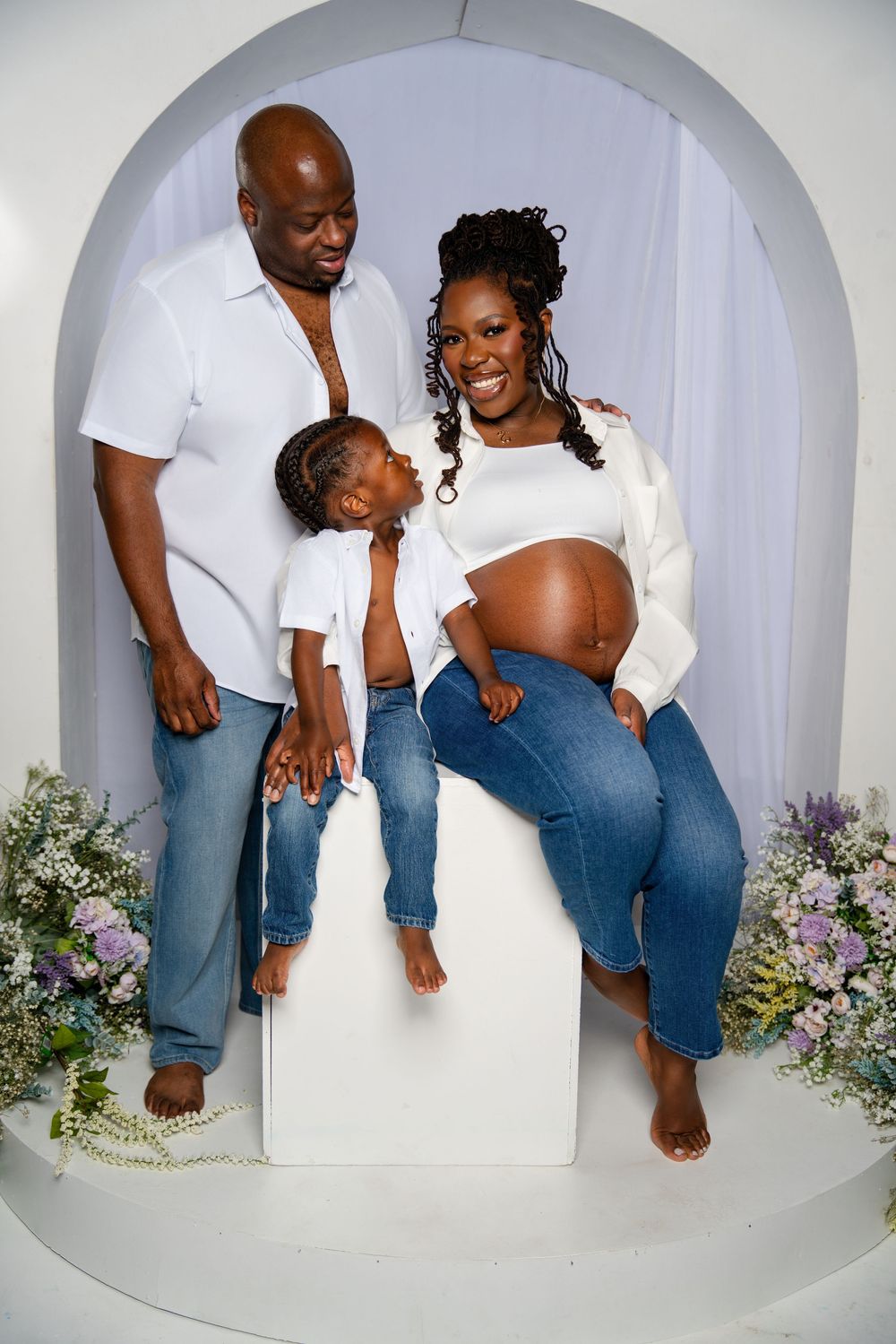 A family maternity photo in a white arch with floral decorations showing a pregnant woman in jeans and a white top.