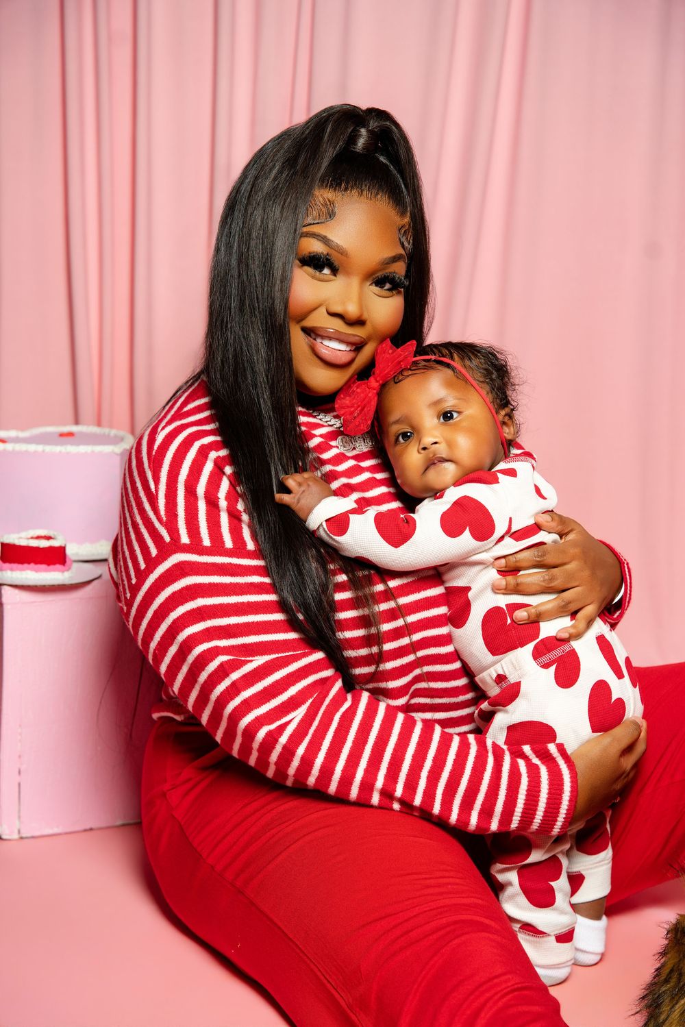A mother and baby wearing matching red and white heart-themed outfits pose together against a pink backdrop.