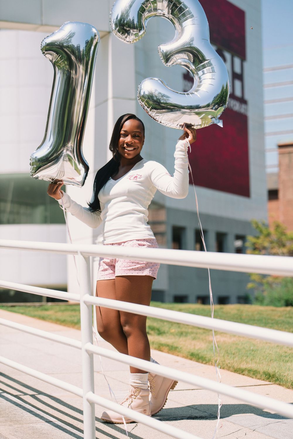 A smiling young woman holds silver number 13 balloons outdoors, wearing a white top and pink shorts.